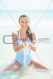 Smiling young woman in swimsuit sitting on sea coast and playing