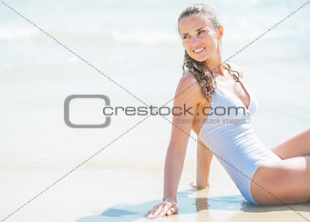 Happy young woman in swimsuit sitting on sea shore and looking o