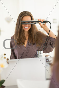 Happy young woman looking through hair straightener in bathroom