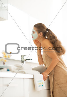 Young woman wearing facial cosmetic mask in bathroom looking in 