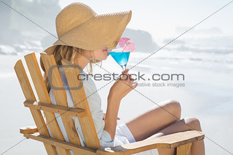 Smiling blonde relaxing in deck chair by the sea sipping cocktail