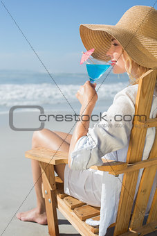 Smiling blonde relaxing in deck chair by the sea sipping cocktail