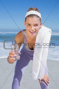 Sporty smiling blonde standing on the beach with towel and bottle