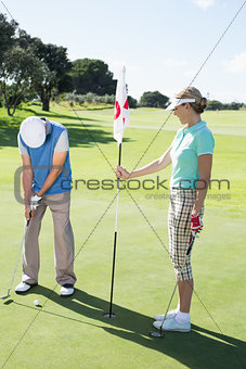 Lady golfer holding eighteenth hole flag for partner putting ball