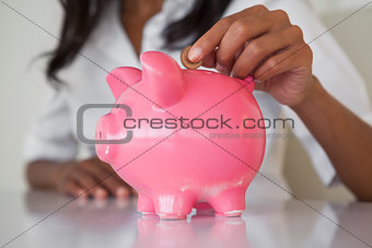 Businesswoman putting coins into pink piggy bank at her desk