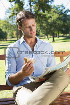 Handsome man sitting on park bench eating sandwich and reading paper