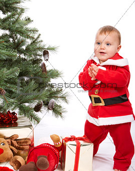 Santa Baby boy standing next to Christmas tree.