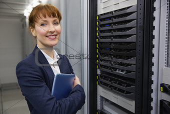 Pretty technician smiling at camera beside server holding tablet pc