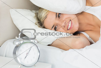 Woman an covering ears with pillow as she looks at alarm clock