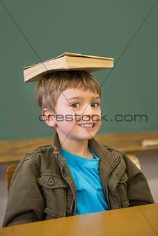 Happy pupil balancing book on his head at desk
