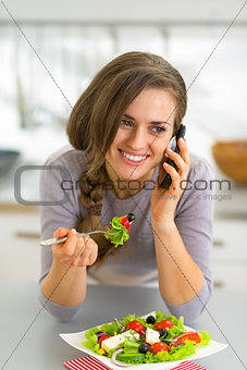 Happy young woman eating greek salad in kitchen and talking cell