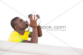 Happy brazilian football fan sitting on couch kissing ball