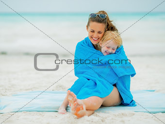 Smiling mother and baby girl wrapped in towel sitting on beach