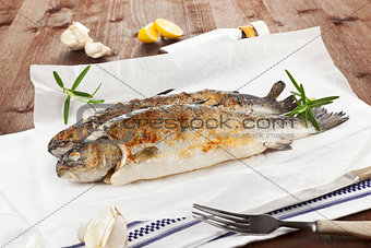 Two grilled trouts with fresh herbs and lemon pieces on wooden t