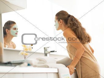 Young woman with facial cosmetic mask looking in mirror in bathr