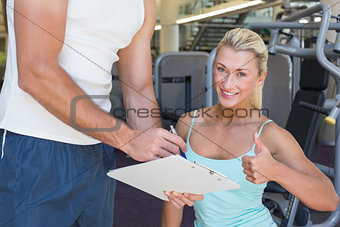 Woman gesturing thumbs up besides trainer with clipboard at gym