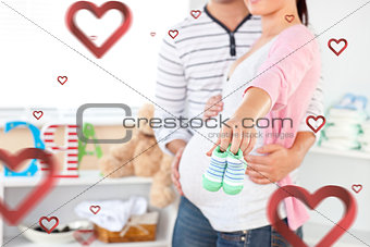 Composite image of close up of a bright pregnant woman holding baby shoes while husband touching her