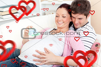 Composite image of portrait of a happy pregnant woman holding baby shoes and of her husband on a sof