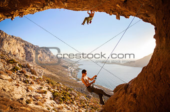 Male rock climber climbing on a roof in a cave, his partner belaying