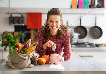 Portrait of happy young housewife putting money into piggy bank 