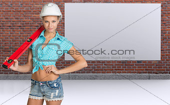 Woman in helmet holding spirit level, smiling. Brick wall and board as backdrop