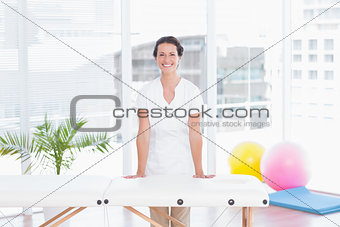 Smiling doctor looking at camera standing behind massage table