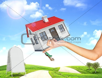 Hand holding house and bunch of keys. In background, green hills with grass, roads, sidewalk sign