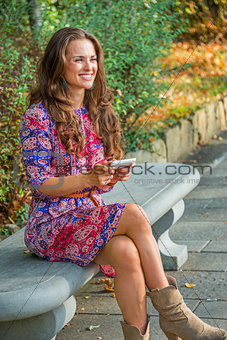 Happy young woman writing sms while sitting on bench in city par