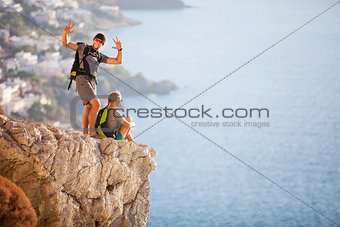 Young couple on rock and enjoying beautiful view, woman sitting on edge, guy posing and making faces