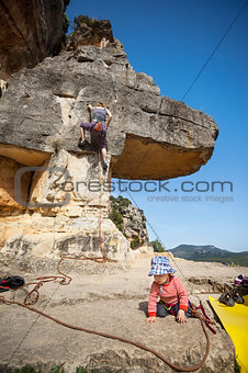 Toddler boy playing at foot of mountain while his mother climbing at the background. The boy is tied in to the rope.