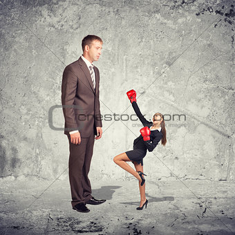 Large businessman looking at small businesswoman in boxing gloves. Concrete wall as backdrop