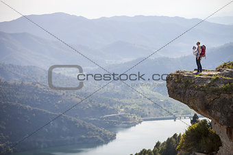 Young mother with baby in sling standing on cliff and enjoying valley view