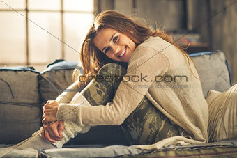Smiling woman on sofa in loft, hugging her knees
