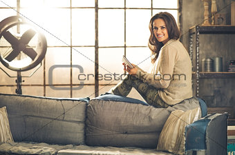 Smiling woman relaxing on sofa back holding a tablet PC in loft