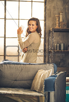 Casually-dressed woman standing in loft by sofa holding phone