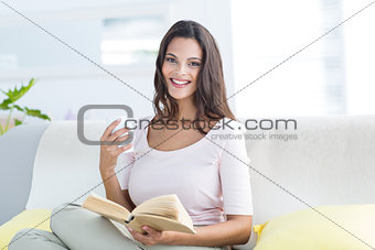 Smiling beautiful brunette holding mug and reading a book while relaxing on the couch