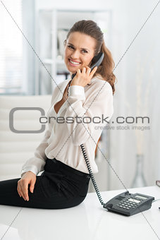 Smiling woman sitting on desk and talking on the telephone