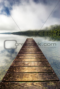 Boat jetty stretching over calm lake with rising mist on the hills, Rotorua, New Zealand