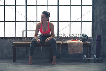Woman sitting on bench in loft gym listening to music on device