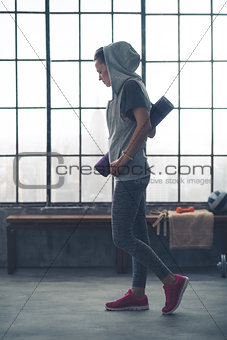 Fit woman wearing hoodie, carrying yoga mat in loft gym