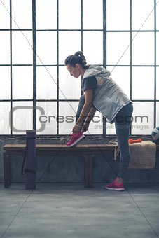 Fit young woman tying shoe on bench by window in city loft gym