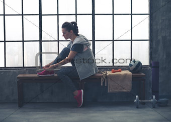 Sporty woman in profile sitting on bench tying shoe in loft gym