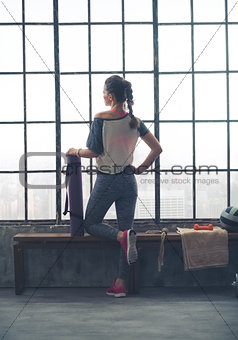 Rear view of fit woman with yoga mat looking out loft gym window