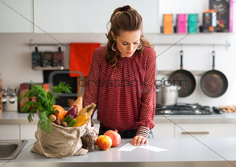 Woman in kitchen reading shopping list on counter with shopping
