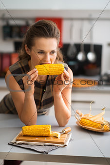 Woman biting into corncob while leaning on kitchen counter