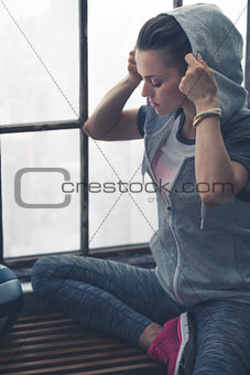 Fit woman putting hoodie on while sitting on loft gym bench