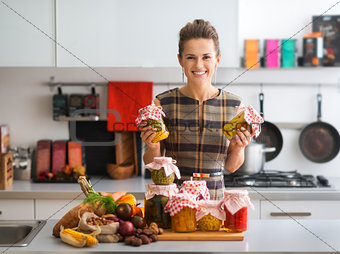 Happy woman in kitchen holding jars of preserved vegetables