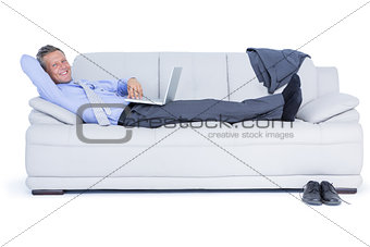 Businessman lying on sofa using his laptop smiling at camera
