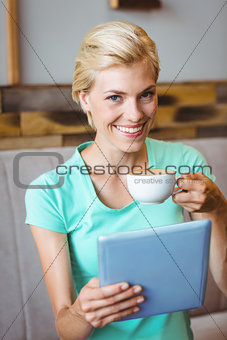 Pretty blonde holding cup of coffee and using tablet computer