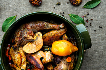 Top view of fried and baked chicken with vegetables in round ceramic stew pot on linen fabric background